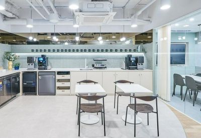 Bright office kitchen area with coffee machines, white cabinetry, and small dining tables.