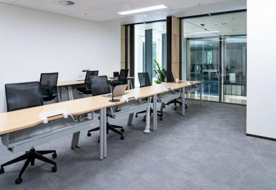 Open-plan office space with rows of light wood desks and ergonomic black chairs.