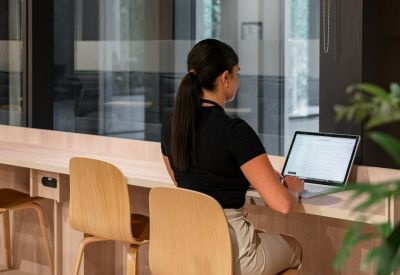 Woman working at a light-filled wooden bench desk overlooking a courtyard through glass walls.