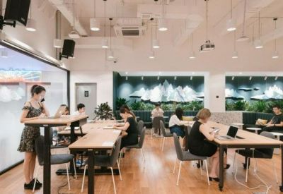 Open-plan coworking space with long wooden tables and a large digital presentation screen.