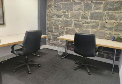 Workstations with light wood desks and black ergonomic chairs against a rustic stone wall.