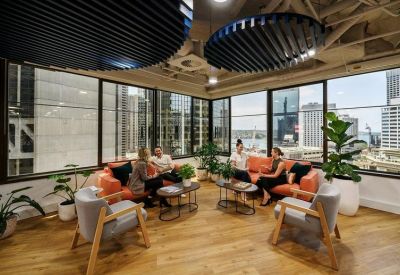 Sunlit lounge area with orange sofas, grey armchairs, and floor-to-ceiling windows.