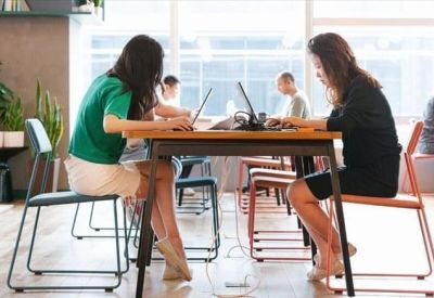 Open-plan workspace where people are working on laptops at a wooden table near large windows.
