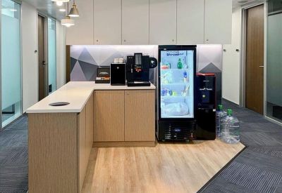 L-shaped refreshment station with a coffee machine, fridge, and wooden flooring.