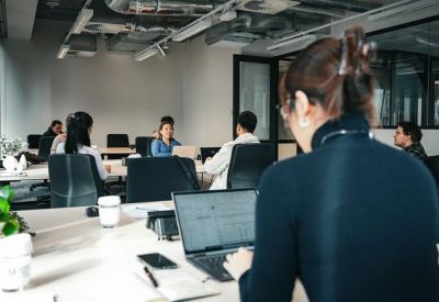 Bright open-plan office with people working at long desks and a plant in the foreground.