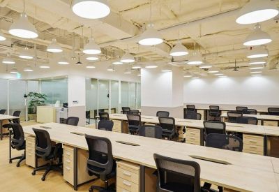Open-plan office with rows of wooden desks, black ergonomic chairs, and industrial ceiling lights.