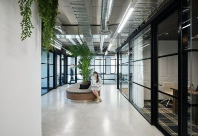Bright corridor featuring glass-partitioned offices, indoor plants, and a curved wooden bench.