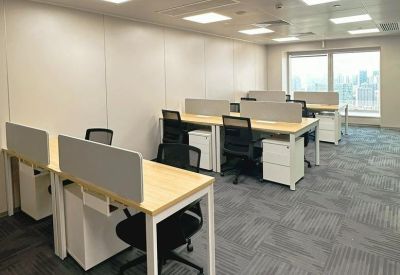 Bright open-plan office space with clusters of wooden desks, white drawers, and grey carpet.