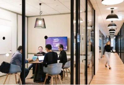 Glass-walled meeting room with people collaborating around a circular table near a bright corridor.