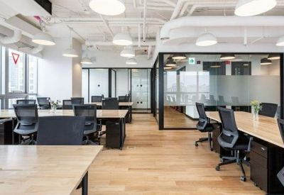Bright open-plan office with long wooden desks, ergonomic black chairs, and glass-walled private suites.