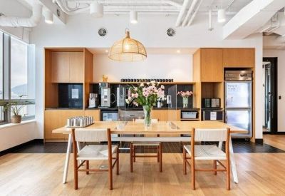Modern communal kitchen with light wood cabinetry, a large dining table, and industrial ceiling details.