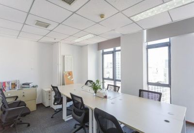 Bright office space featuring long white shared desks, black chairs, and large windows with city views.