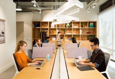 Open-plan office space with individuals working at long wooden desks under geometric lighting.