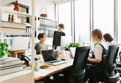 Bright open-plan office with people working at long wooden desks by large windows.