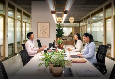 Open-plan office space with long white desks and people working at laptops.