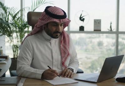 Man in traditional attire working at a wooden desk with a laptop and city view.