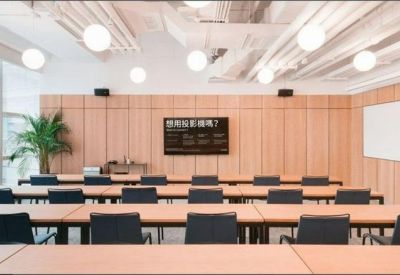 Professional training room with rows of wooden desks, black chairs, and a wall-mounted screen.