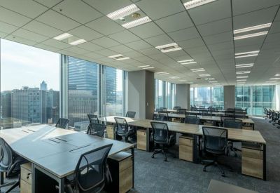 Open-plan office with rows of wooden desks and ergonomic chairs near large windows.