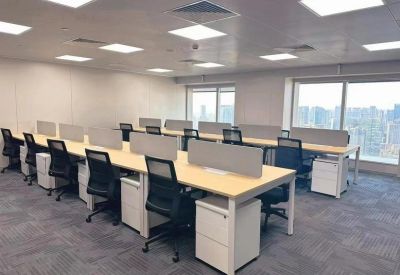 Open-plan workspace with long rows of desks, privacy dividers, and city views through large windows.