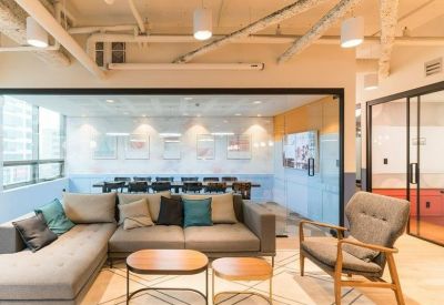 Lounge area with a grey L-shaped sofa, wooden coffee tables, and a glass-enclosed meeting room behind.