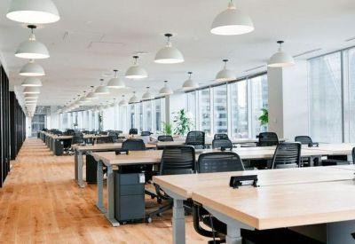 Modern open-plan office with rows of wooden desks, black ergonomic chairs, and large windows.