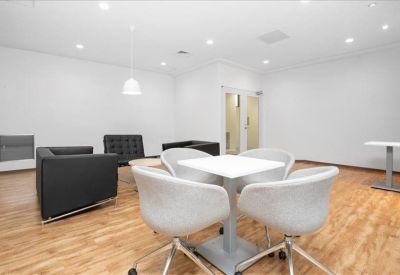 Bright communal lounge featuring white armchairs around a small table and a black sofa.