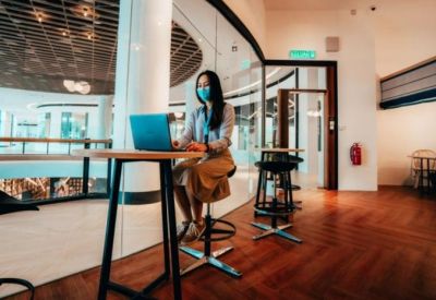 Coworking area with high tables and stools where a person in a mask works on a laptop near large glass windows.
