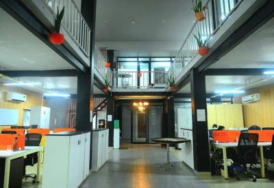 Spacious two-level industrial coworking space with mezzanine walkways, plants, and orange office chairs.