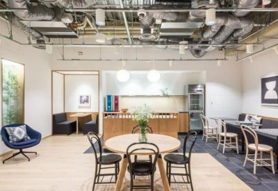 Bright communal dining area with round wooden tables, black chairs, and exposed ceiling pipes.