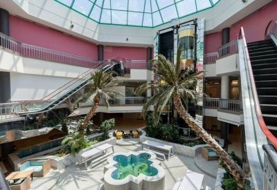 Indoor courtyard area with palm trees, white benches, and a central water feature under a glass ceiling.