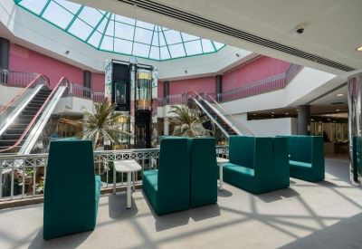 Bright atrium lounge with teal high-back booths, escalators, and a large geometric skylight.