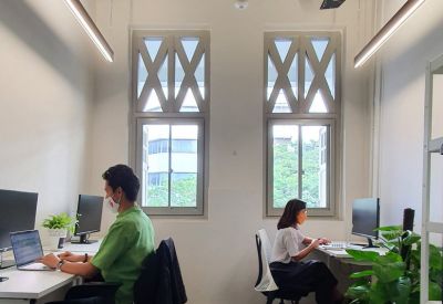 Bright shared office space with two desks, computers, and large traditional windows.