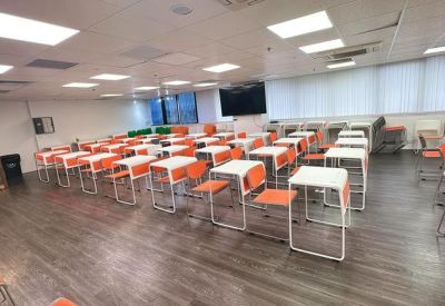 Wide-angle view of a seminar room with orange seating, white tables, and a large wall-mounted screen.