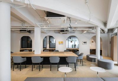 Communal wooden work table surrounded by grey armchairs and arched windows.