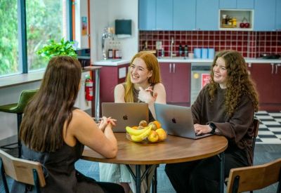 Three people collaborating at a round wooden table in a bright communal kitchen and breakout area.