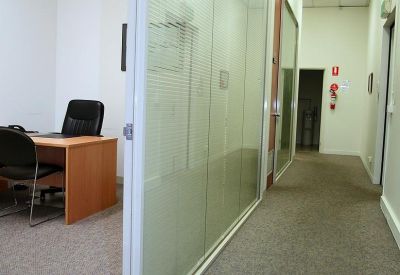 Private office with a light wood desk and black leather chair by a glass partition.