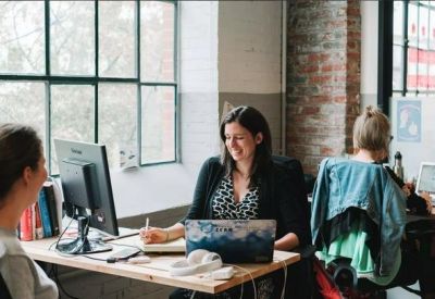 Open-plan office with employees working at wooden desks near large windows.