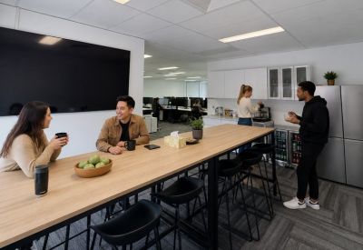 Bright communal kitchen and breakout area with a long timber table, black bar stools, and a fruit bowl.