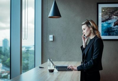 A professional woman working at a standing desk with a laptop against a large window with city views.