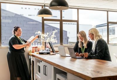 Bright communal kitchen area with a large wooden island, laptop users, and city views.