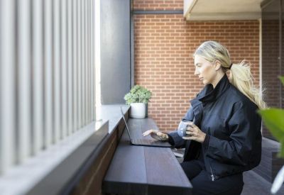 Woman working on a laptop at a wooden balcony bar with a brick wall background.