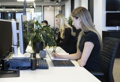 Two women working at modern white desks with indoor plants and large monitors.