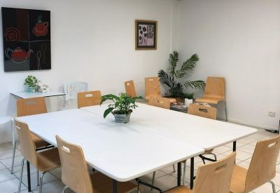 Communal breakroom with a large white square table, wooden chairs, and potted plants.