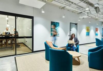 Modern breakout lounge with blue armchairs, a glass meeting pod, and exposed ceiling pipes.