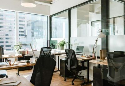 Open-plan workspace with wooden desks and black ergonomic chairs near large windows.