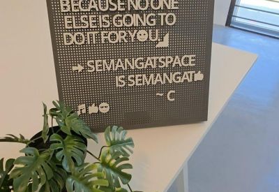 Inspirational signage on a grey pegboard placed next to a Monstera plant.