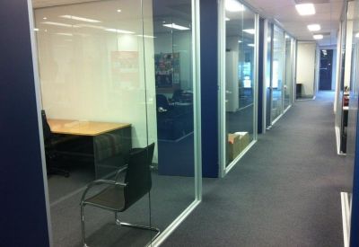Office corridor showing glass-partitioned workspaces and grey carpeting.