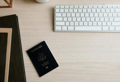 Close-up of a wooden desk with a white keyboard, a black passport, and a notebook.