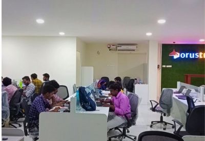 Open-plan office with people working at desks and a green moss wall featuring a neon sign.