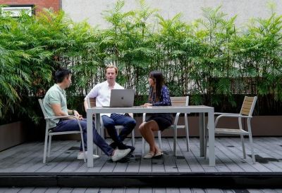 Professional team meeting at a grey table on an outdoor terrace lined with greenery.